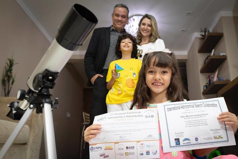 Em primeiro plano, menina de 6 anos sorri e mostra certificados orgulhosamente. Atrás, homem adulto de terno, mulher adulta de cabelo loiro e roupa branca e menino de 8 anos de blusa amarela